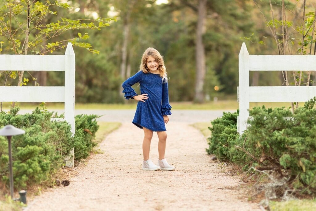 A little girl in a blue dress stands between two sections of white picket fence