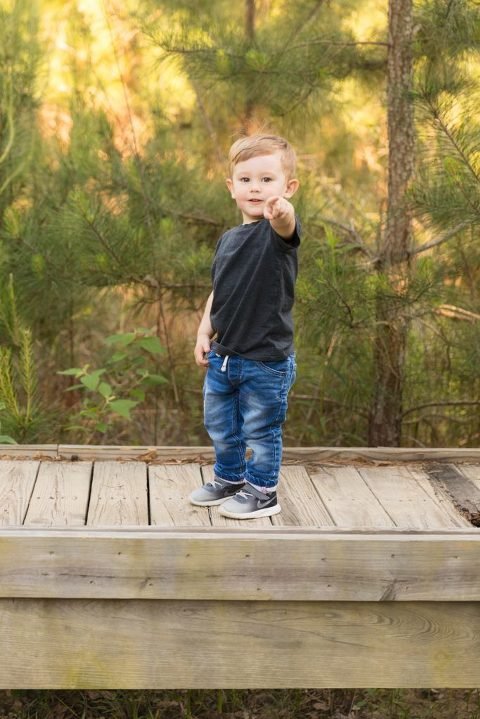 Woodlands Child Photographer - Little boy stand on a bridge and points towards the camera