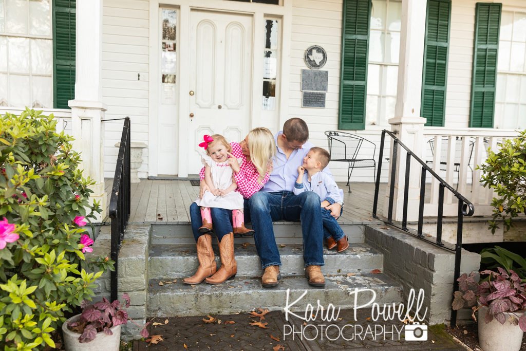 A family of four sits on the steps of an white historic home with green shutters. 