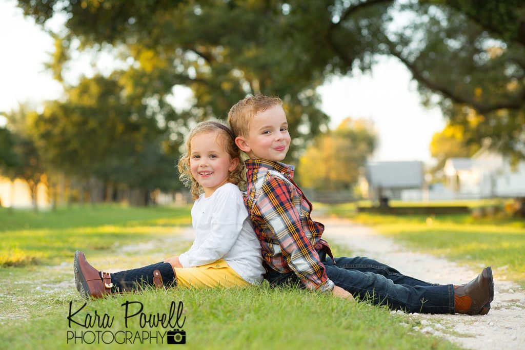 Two kids sit back to back on a grass covered lane posing for the woodlands tx photographer