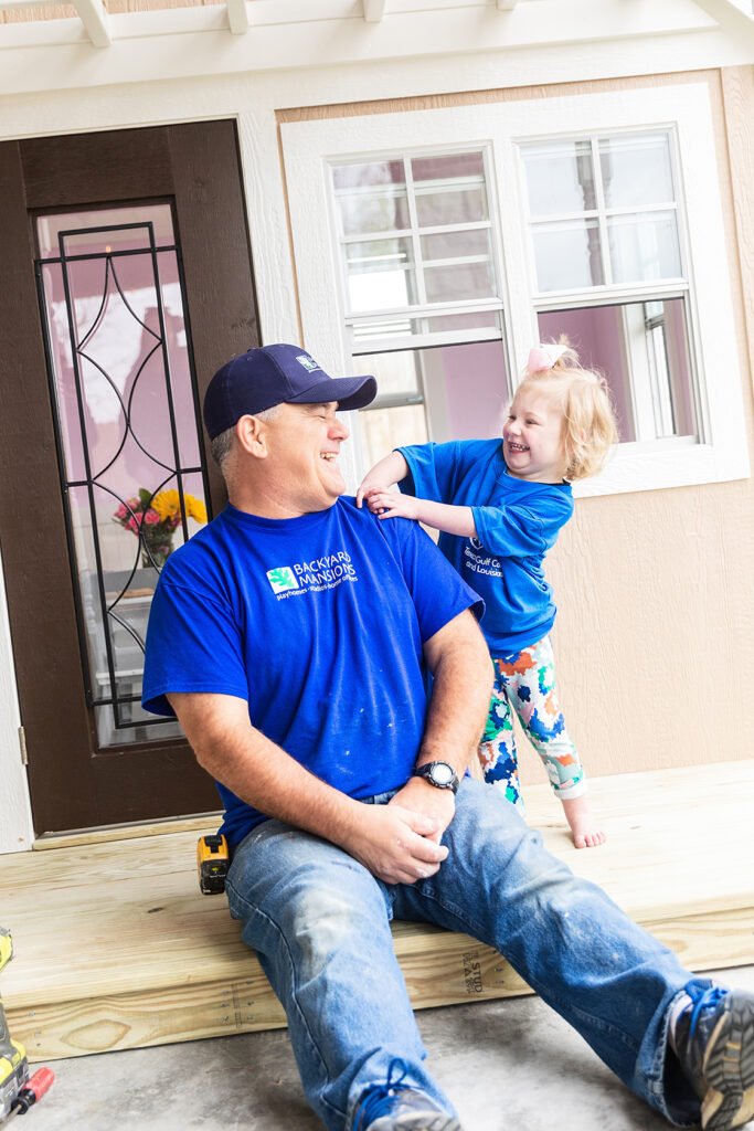 A child stand behind a man who is sitting on the ground. Both are laughing