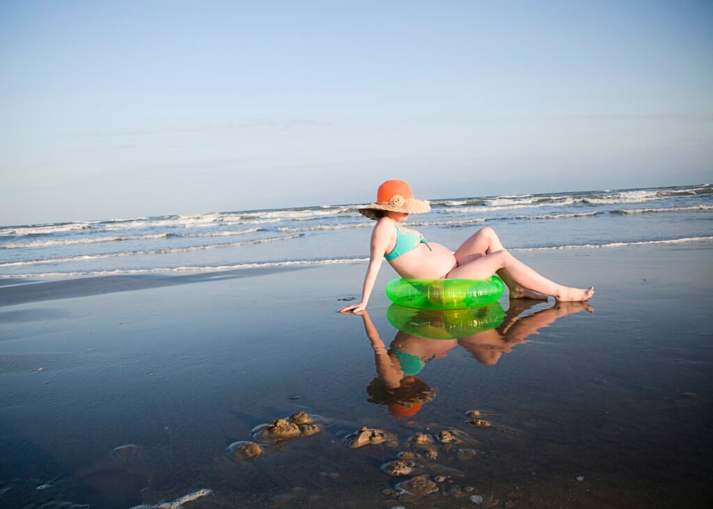 Maternity photo at the beach in Galveston. A woman is sitting in a green floatie getting sandy toes photos