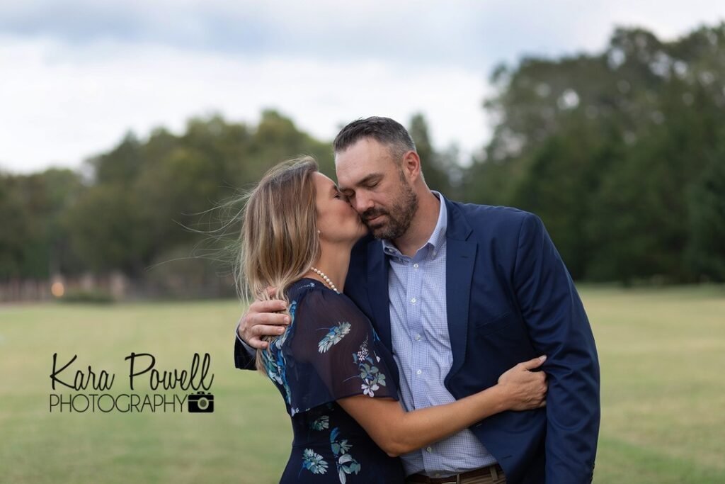 A husband and wife hug and she kisses his cheek during a fun photo session.