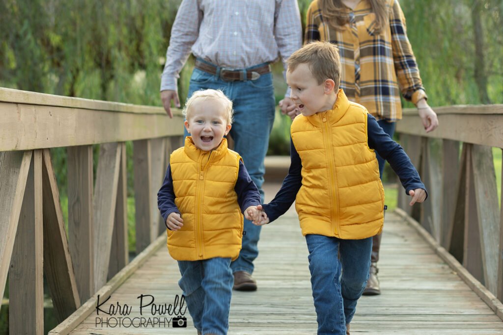 Two brothers run across a wood foot bridge with their parents following behind. Captured by The Woodlands Brother Photographer