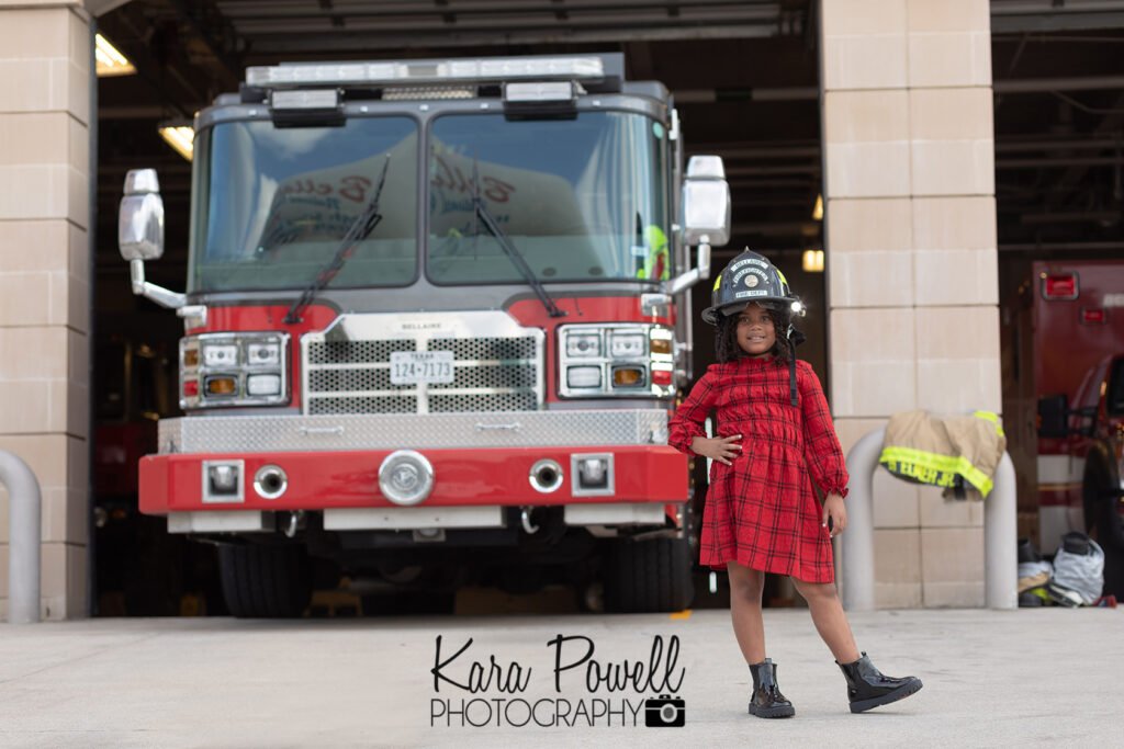 Little girl standing in front of a fire truck and a fire station doing her very own firefighter photo session