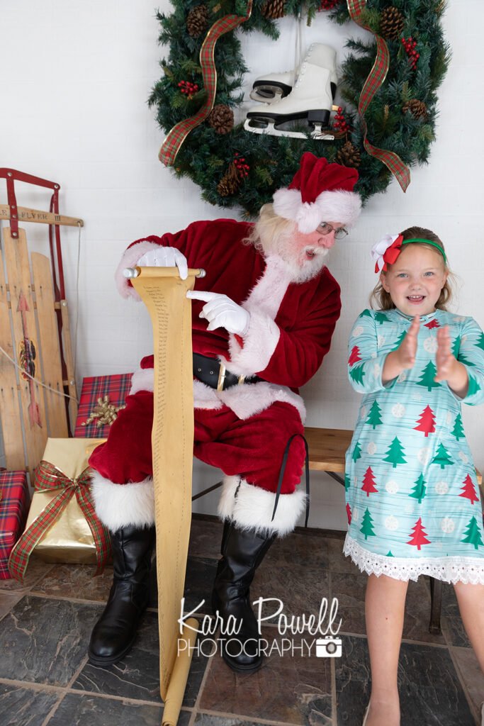 A little girl sits with Santa Claus and claps because she is on the nice list at her Quiet Time with Santa session.