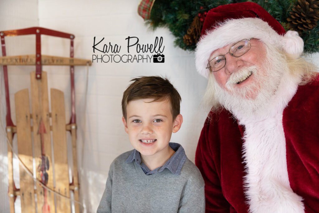 A little boy smiles with Santa and a classic snow sled at his Quiet Time with Santa session.