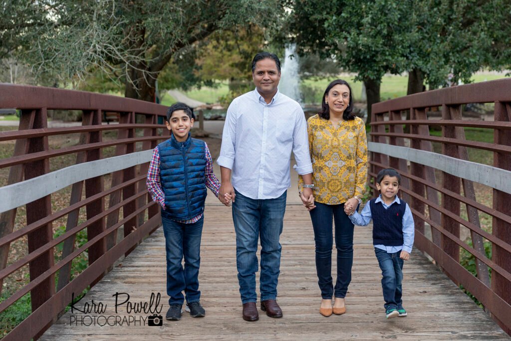 A family of four stand on a wooden foot bridge with metal railings. Everyone even the 3 year old is smiling!