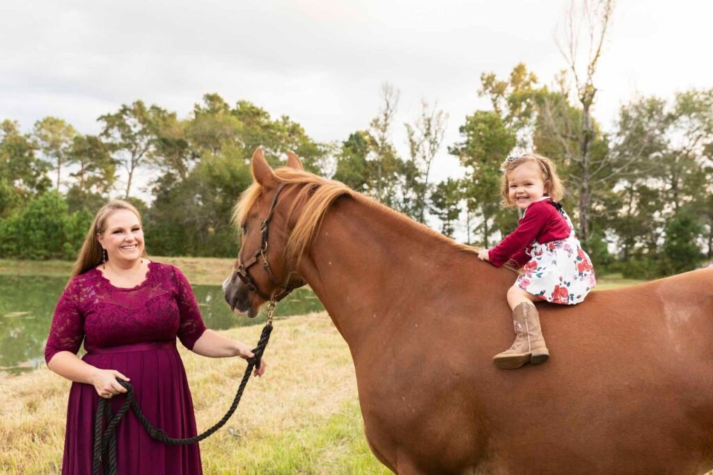 A little girl in the Tippy Toes Baby Plan sits atop her horse, led by her mother.