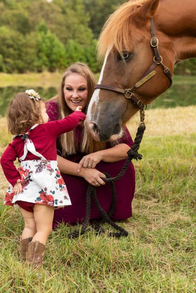 A little girl who is in the toppy toes baby plan pets the nose of her horse while her mother looks on.