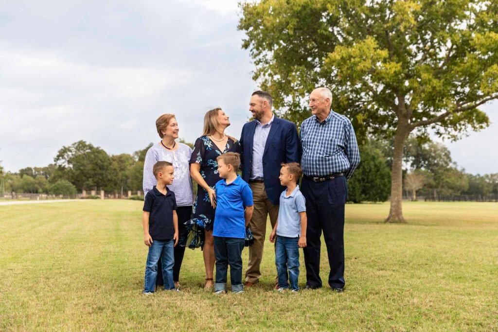 Extended family of 7 stands in front of a large tree in a field.