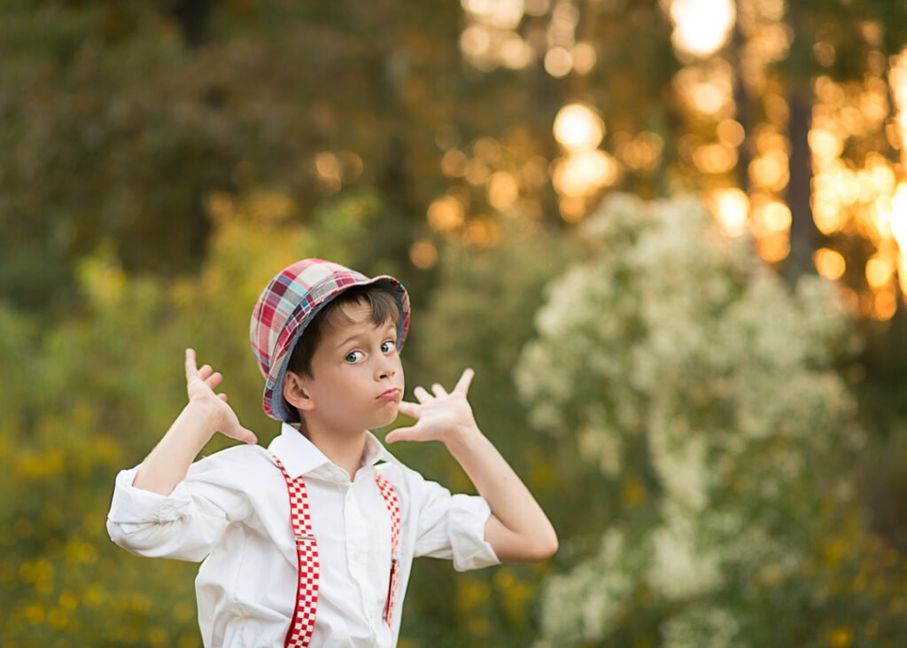 A little boy wears a hat and suspenders and poses with a silly face for a photographer of kids.