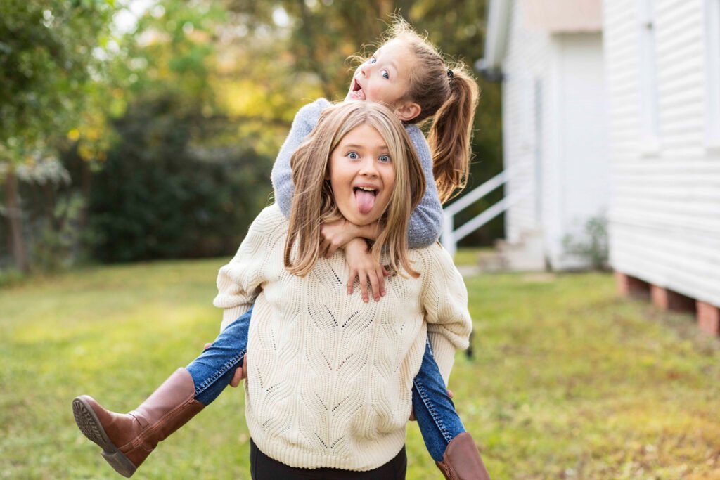 An older sister holds her younger sister in a piggy back while they make silly faces at their silly family photographer