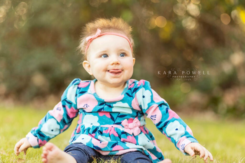A 9 month old little girl with short spiky hair sits in the trees in preparation for her 1st birthday session.