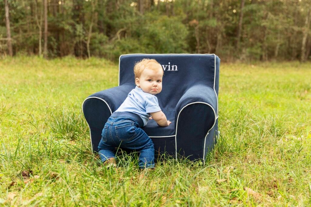 One year old boy frowns and looks over his shoulder while standing in front of a blue chair at his one year photo session.