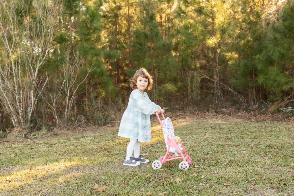 A little girl pushes her baby stroller through the woods while taking family portraits in the woodlands